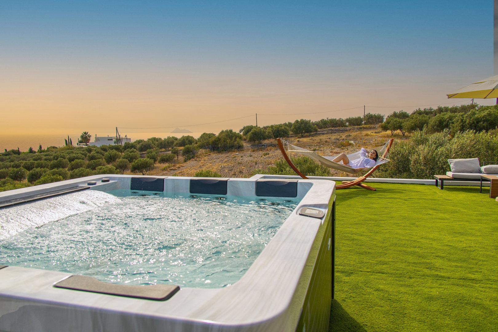 A hot tub on a grassy patio overlooks a landscape of olive trees and the sea at sunset, with a woman relaxing in a hammock in the background.