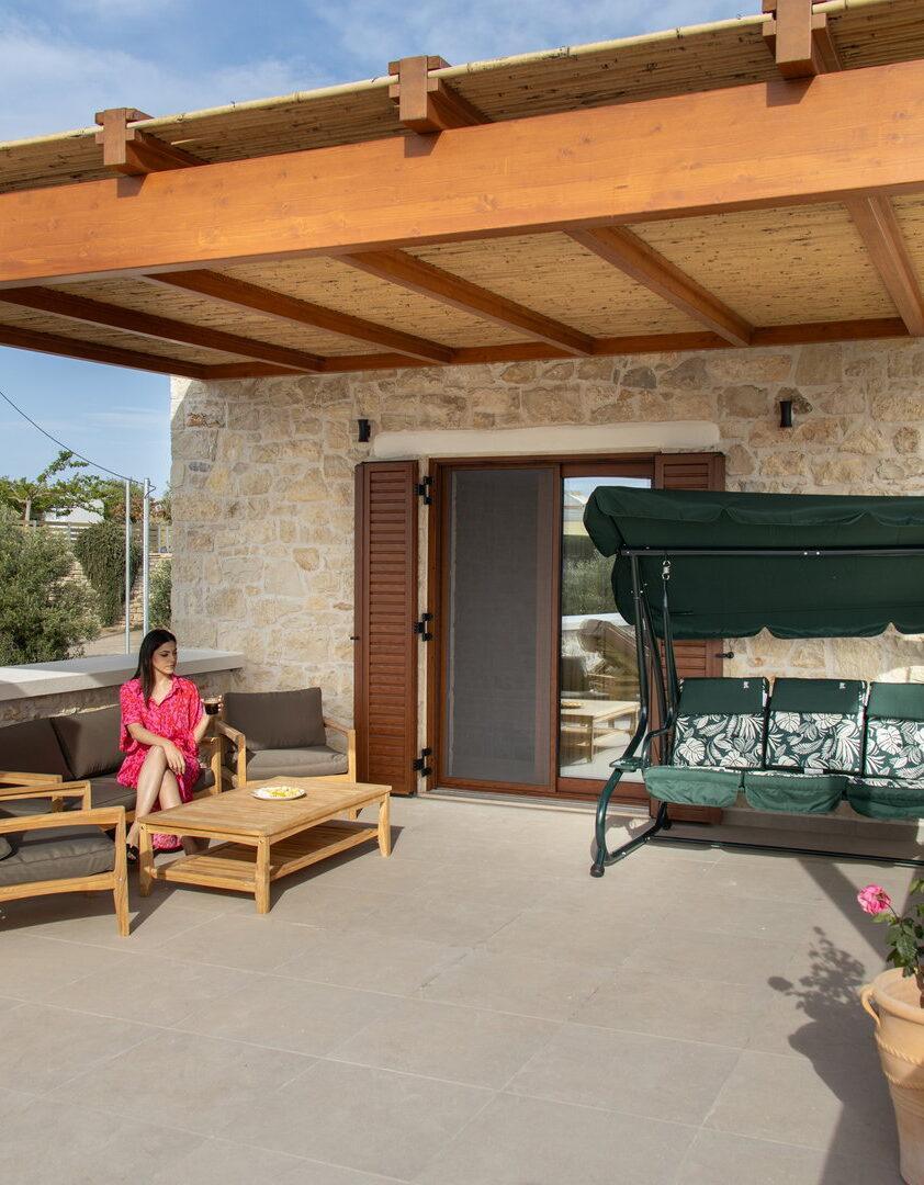 A woman in a pink dress sits on an outdoor sofa under a wooden pergola, next to a matching coffee table and a green porch swing, with a stone house in the background.